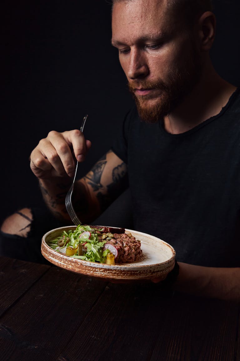 A bearded man enjoys a gourmet meal indoors, showcasing delicious meat and salads.