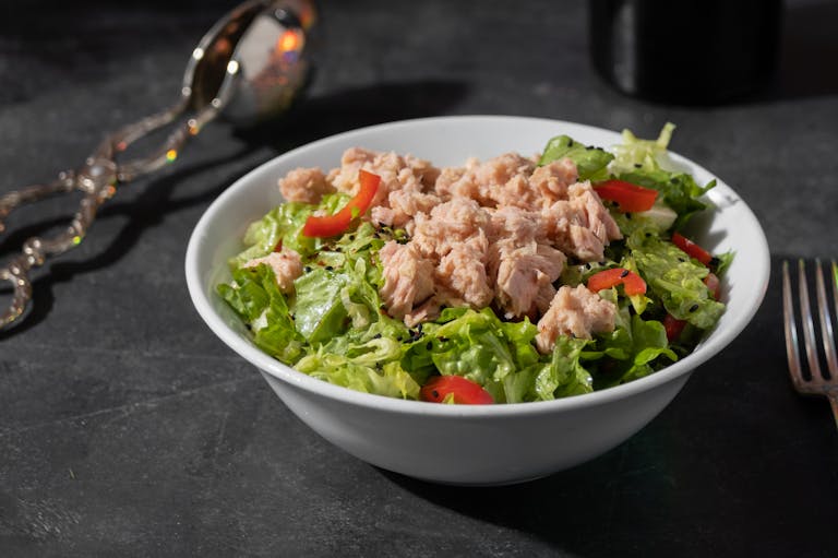 A close-up of a fresh tuna salad featuring lettuce and tomatoes in a white bowl on a dark surface.