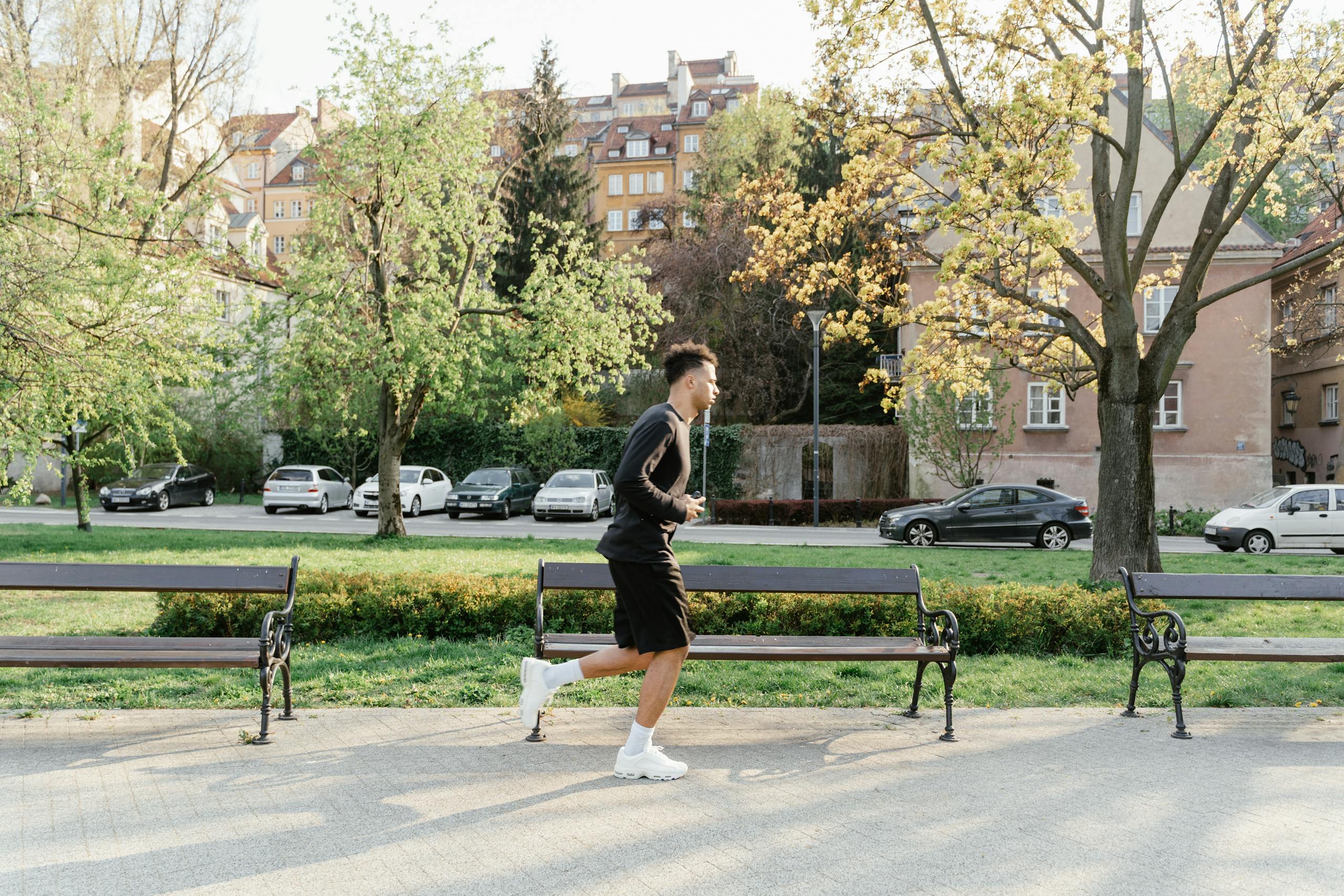 A man in athletic wear jogging through a sunlit park. Urban backdrop with trees and benches.