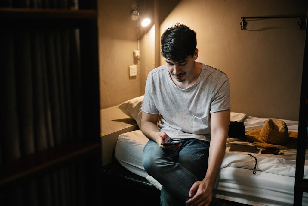 A young man sits on a bed, browsing on his smartphone in a cozy bedroom atmosphere.