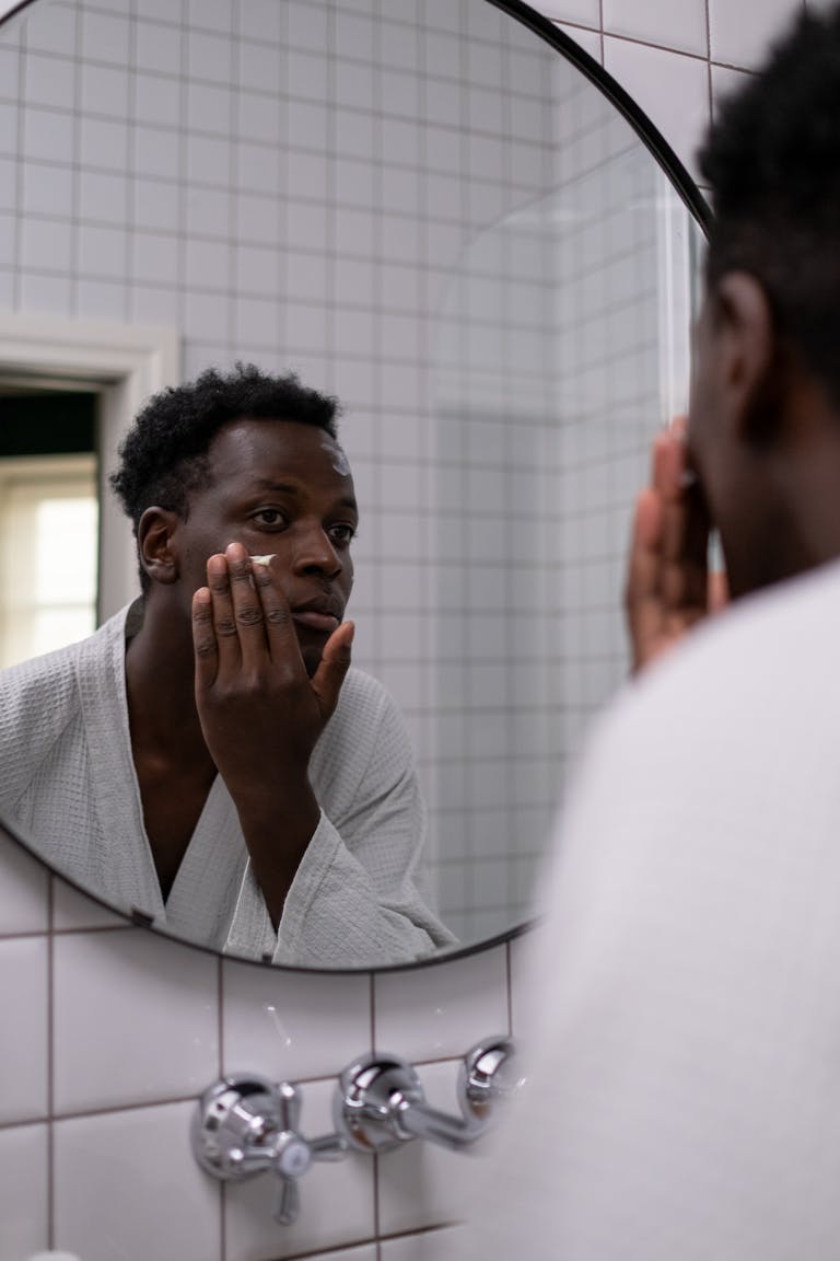 African American man applying facial cream while looking in a bathroom mirror, promoting skincare routine.