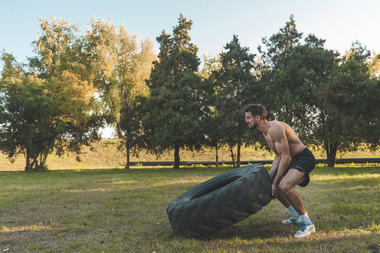 Athletic man flipping large tire during outdoor workout in sunny park setting.