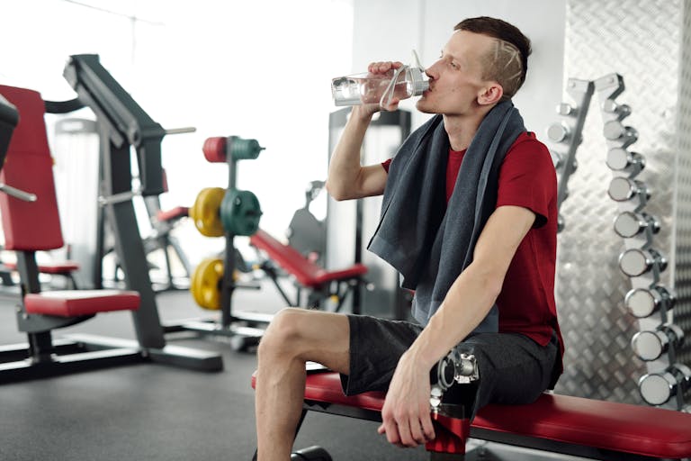 Man with prosthetic leg rests on bench and drinks water during gym workout.