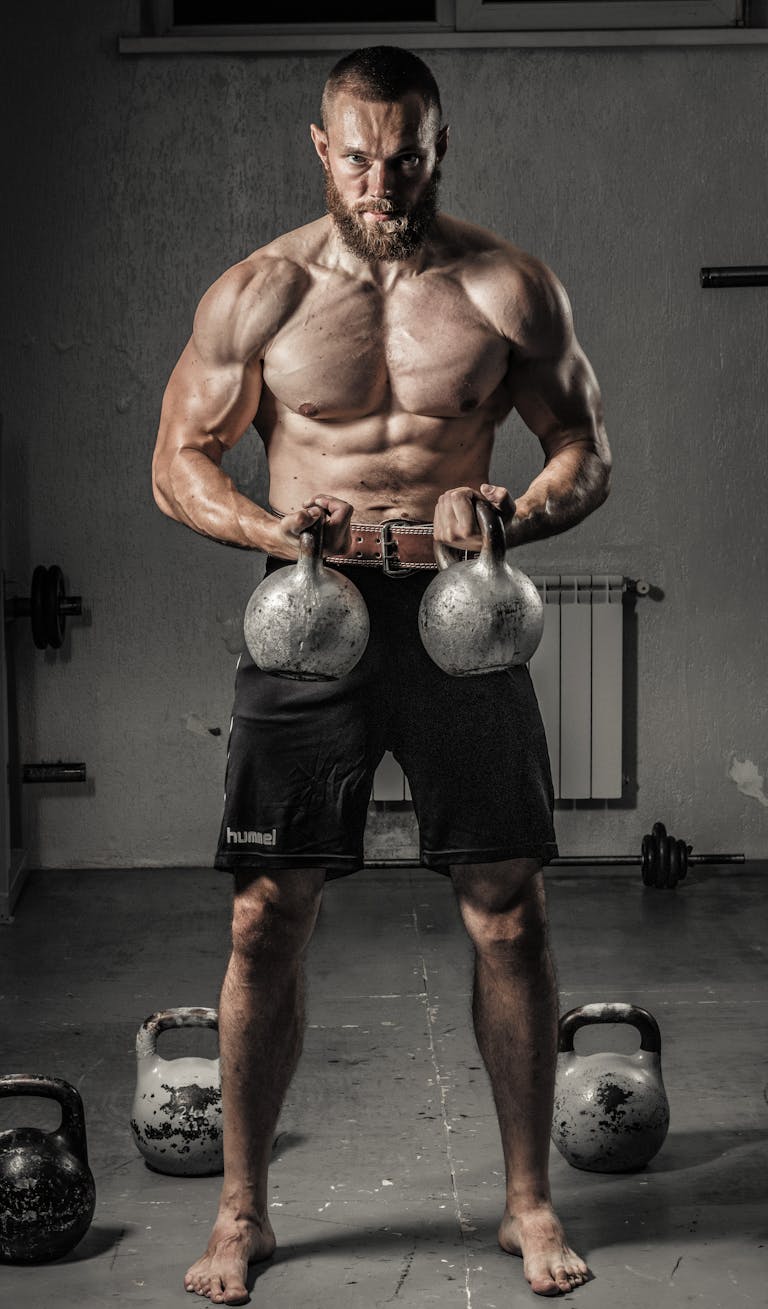 Shirtless man with muscular build lifting kettlebells indoors, showcasing strength and fitness.